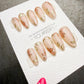 A close-up of a set of pink nail art designs featuring gold leaf patterns, displayed on a white background.