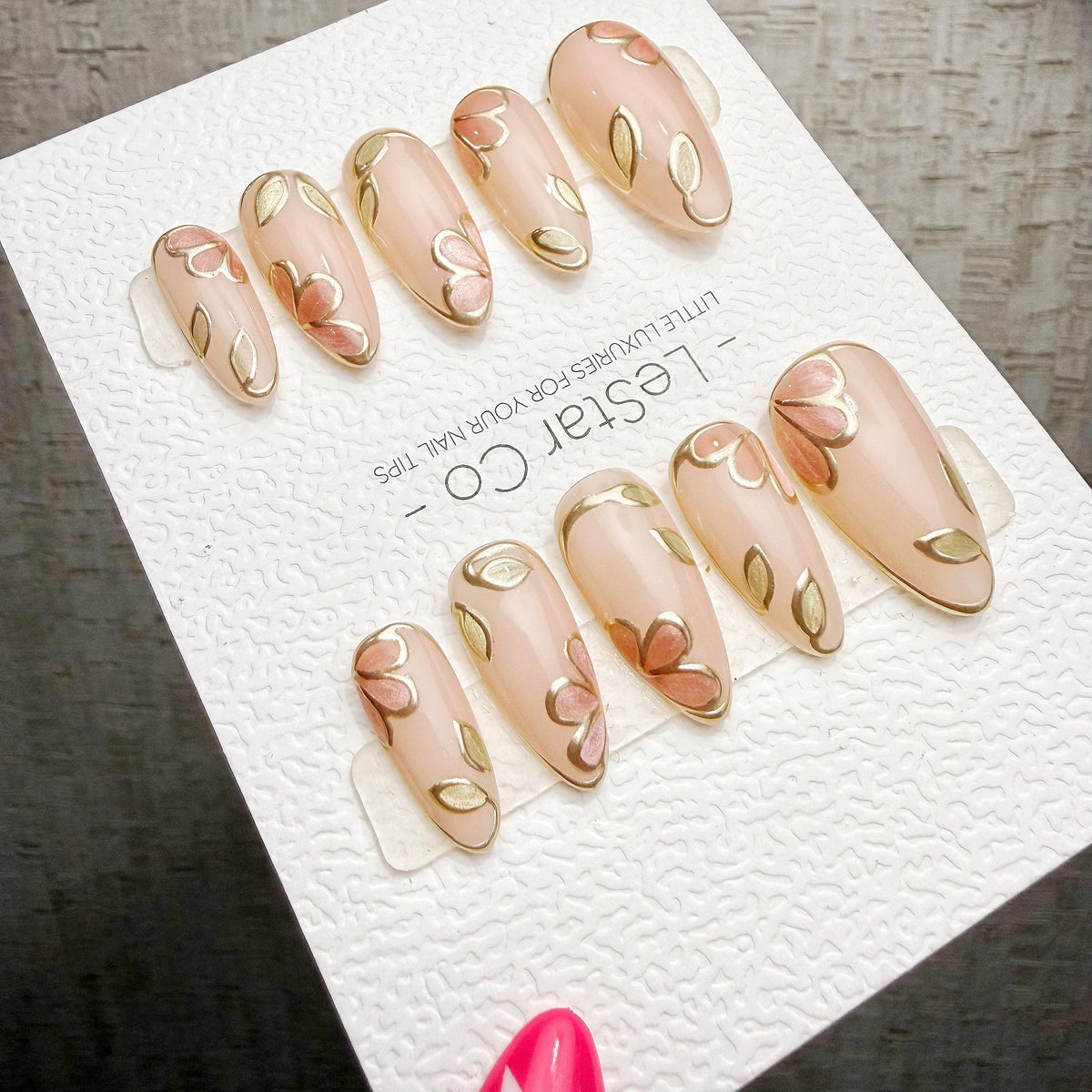 A close-up of a set of pink nail art designs featuring gold leaf patterns, displayed on a white background.
