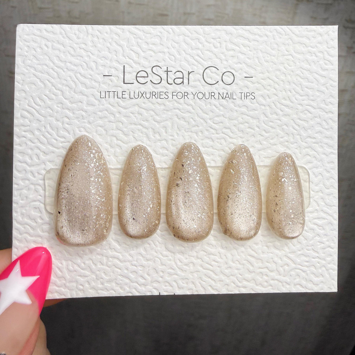 A set of gold glittery nail tips is displayed on a white background, with a hand holding the product.